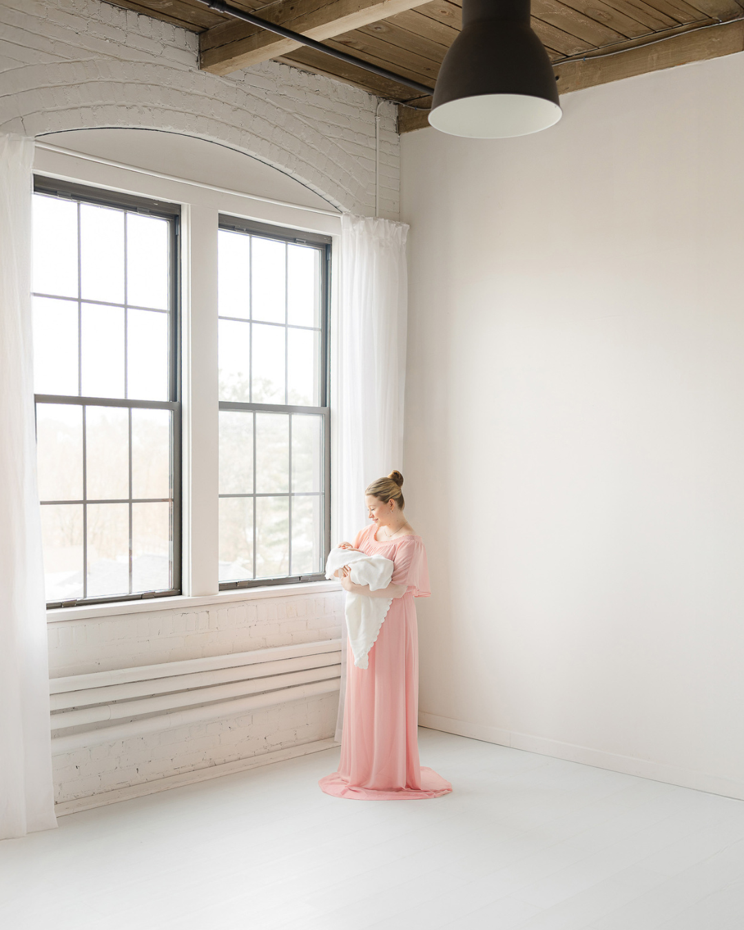 Mother holding her newborn by a large window in a light-filled Boston photography studio with tall ceilings and soft natural light.