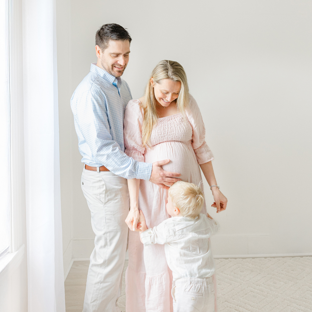 pregnant mom wearing boston maternity clothes stands with husband smiling down at toddler during photoshoot
