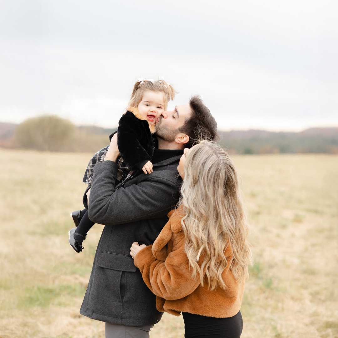 Central MA family photographer captures joyful fall moment of dad kissing daughter’s cheek while mom looks on with love in an open field