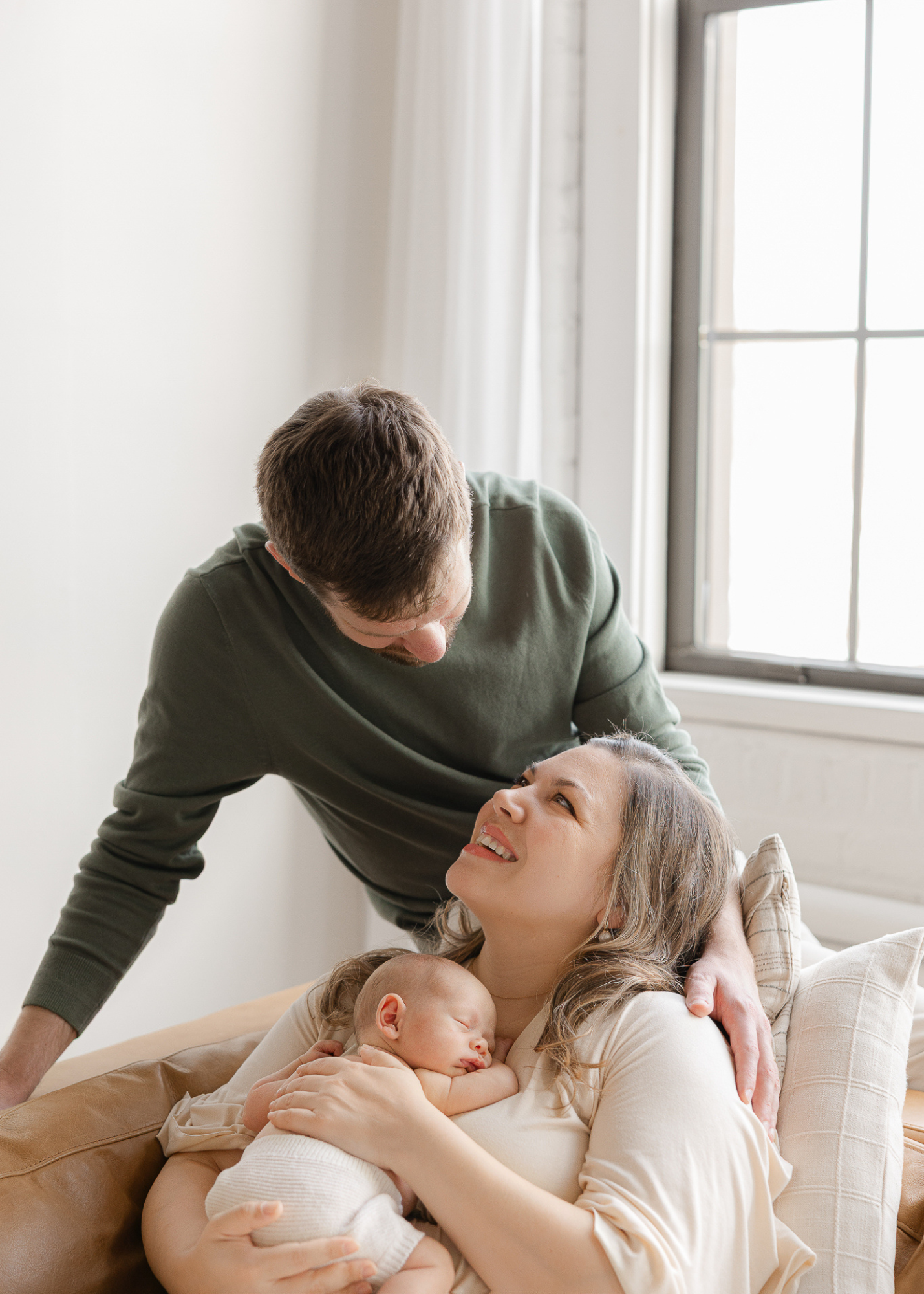 Lifestyle newborn family portrait of dad leaning in while mom cuddles their sleeping newborn baby on a cozy couch, photographed by a Massachusetts newborn photographer.