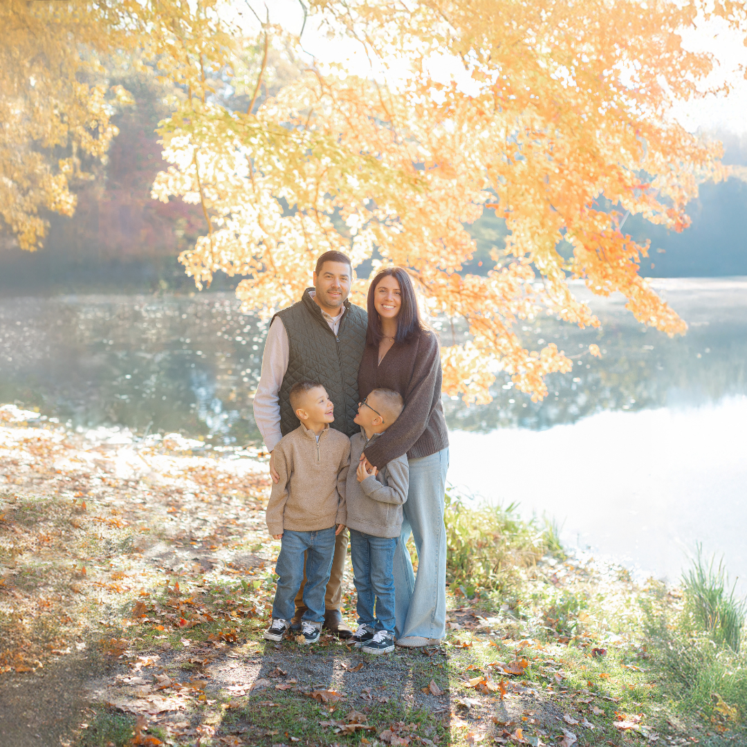 Family of four standing together beneath colorful fall foliage during a family photography session at Choate Park in Medway, MA.
